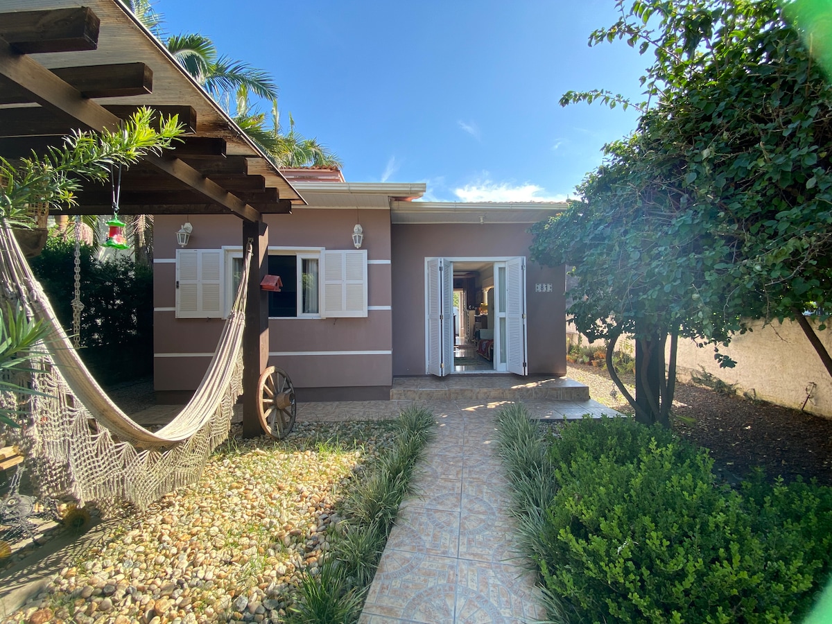 A welcoming entrance is featured, with a pathway leading to an open door. Lush greenery surrounds the house, and a hammock is seen suspended nearby. The exterior has light-colored walls with decorative shutters and is complemented by a bright blue sky above.