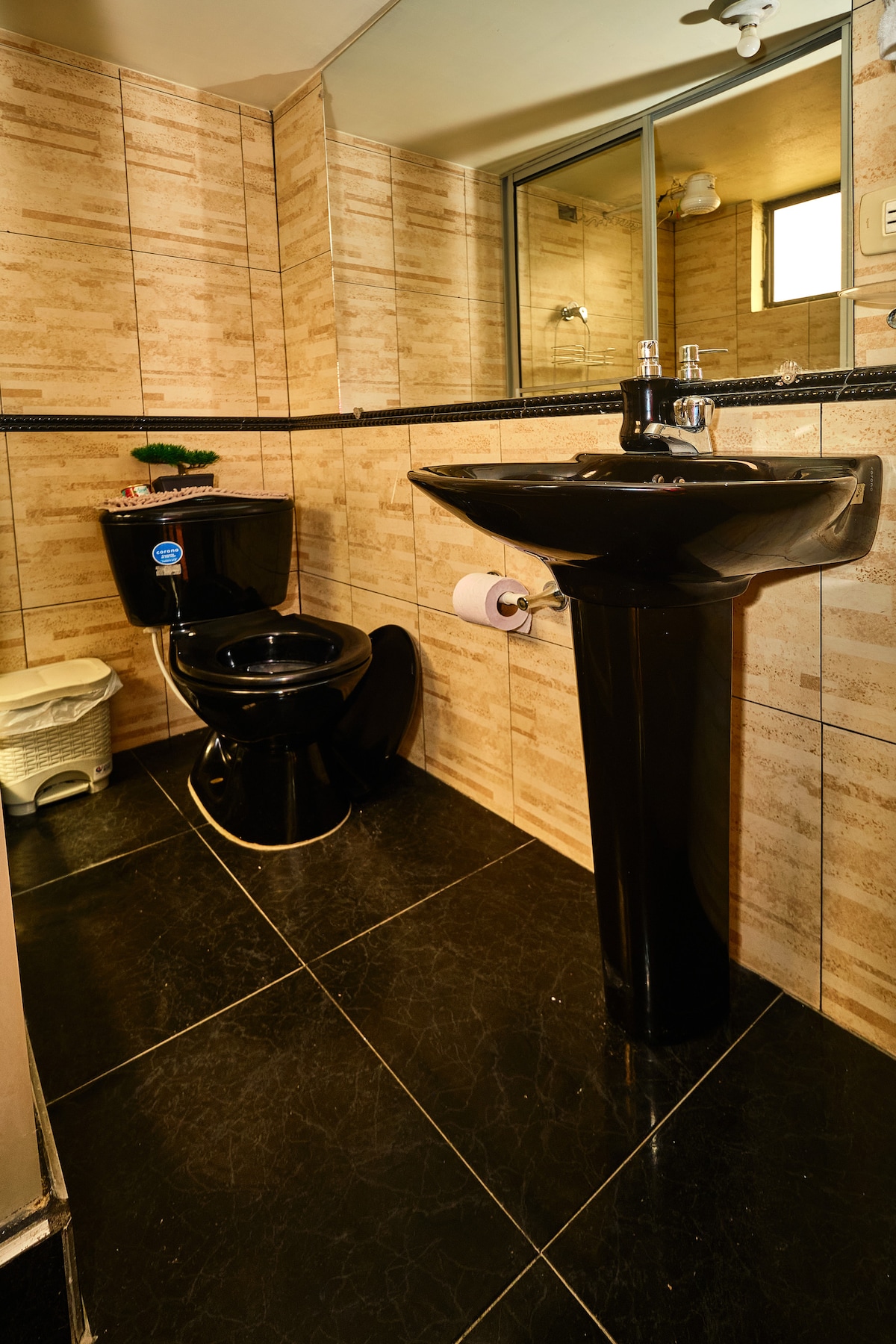 A compact bathroom features a black toilet and matching pedestal sink against a backdrop of textured beige wall tiles. A small wastebasket is visible near the toilet. A mirrored wall reflects the light and enhances the sense of space.