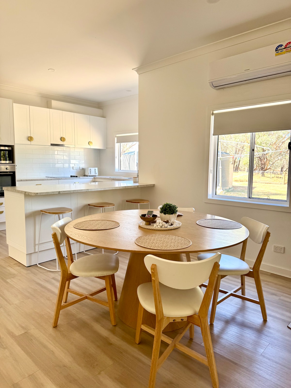 A well-lit dining area features a round wooden table surrounded by four white chairs. The kitchen is equipped with modern appliances, including a built-in oven and stovetop, and is complemented by bright, airy windows that provide views of the surrounding yard.
