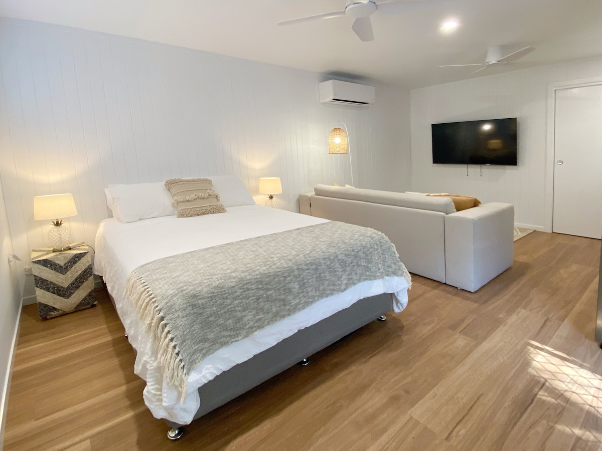 A cozy bedsit style accommodation features a spacious bed dressed in neutral tones. A seating area with a light-colored sofa is positioned nearby, complemented by soft lighting from bedside lamps. Natural wood flooring extends throughout the room, creating a warm ambiance.