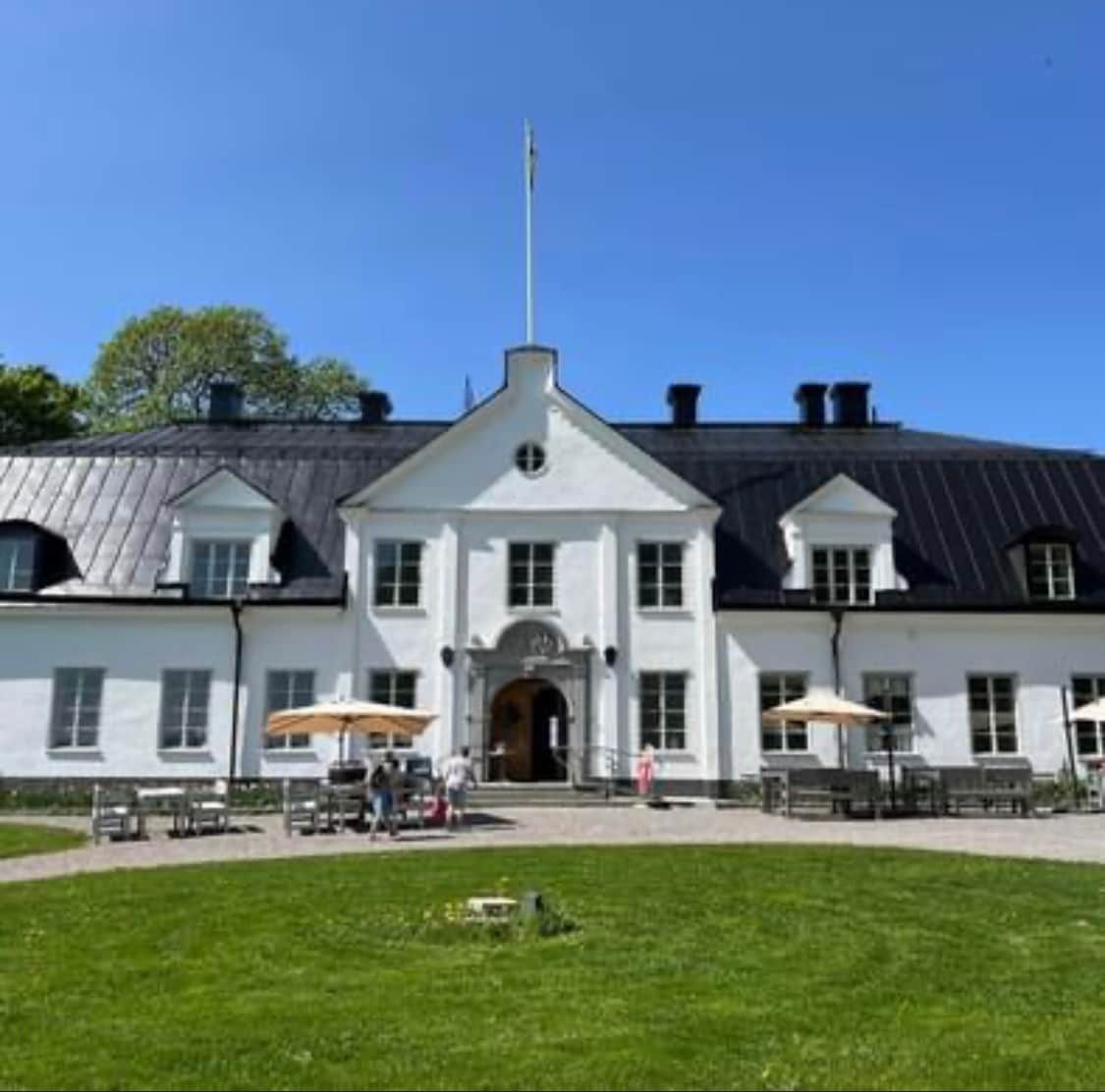 A classic white manor house is set against a clear blue sky. Large umbrellas provide shade over wooden tables and chairs arranged on a green lawn, inviting outdoor gatherings. The architecture features a symmetrical design with a prominent entrance, enhancing the tranquil atmosphere of the grounds.