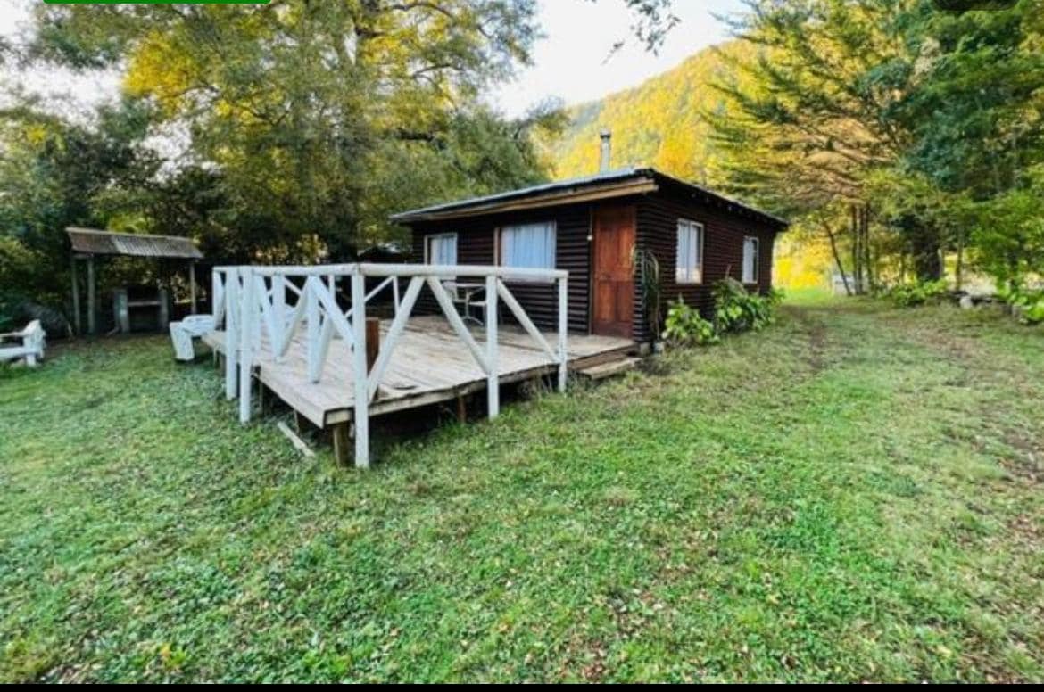 A rustic cabin is set among green foliage, featuring a wooden deck with railings that wrap around. Large windows are visible, allowing natural light to enter. The surrounding grassy area provides a tranquil and serene atmosphere, complemented by distant trees and a gentle landscape.