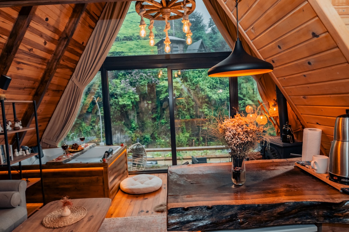 The interior features a wood-paneled design with an angled ceiling and large glass windows that showcase a scenic outdoor view. A modern light fixture hangs above a wooden table, while a cozy seating area with a soft rug and decorative items is visible.