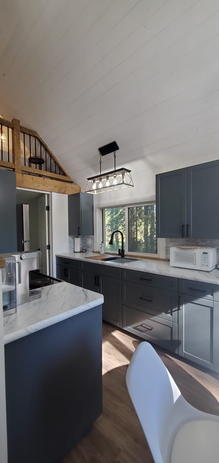 A modern kitchen is featured with dark cabinetry and a light marble countertop. Natural light enters through a large window, illuminating the space. A dining table is visible, along with a microwave and a stove, highlighting the functional layout of the area.