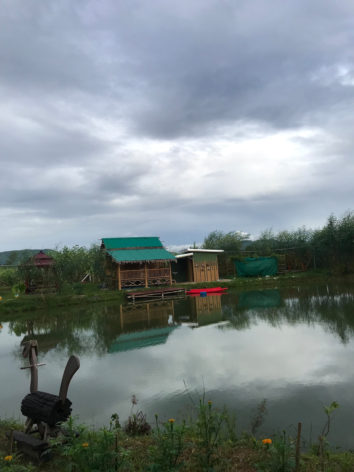 A rustic wooden structure with a green roof is reflected in the calm waters of a pond, surrounded by lush greenery. A nearby shed features a light-colored exterior. Overcast skies create a serene atmosphere in the landscape.