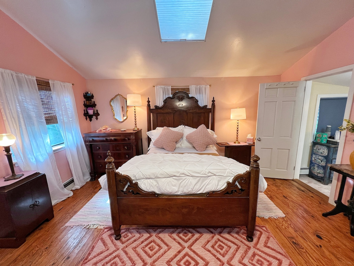 A spacious bedroom features a large wooden bed with decorative headboard and soft white bedding. Two bedside tables with lamps are positioned on either side, complemented by a wooden dresser. Natural light enters through a skylight, and pale pink walls enhance the serene atmosphere.