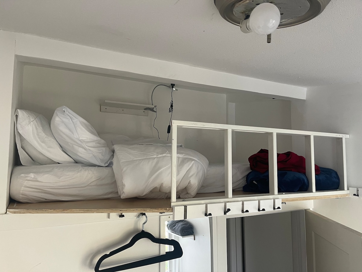 A lofted sleeping area displays a neatly made bed with fluffy white pillows and bedding. A small shelf above the bed offers additional storage. Clothes are neatly folded and placed nearby, while a clothes hanger is attached to the wall below.