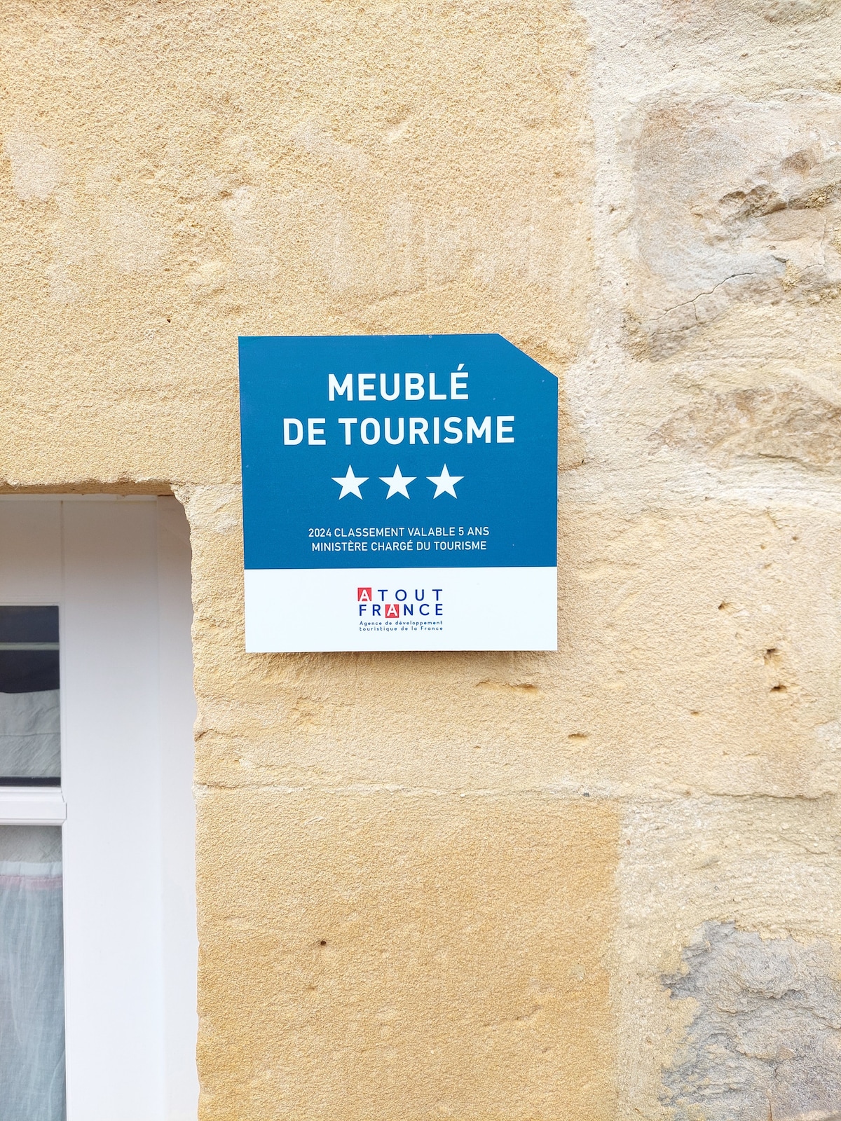 A tourism classification sign affixed to a textured stone wall is displayed prominently. The sign indicates a rating of three stars and contains text in French, showcasing the property's official recognition. A glimpse of a nearby window is visible in the background.