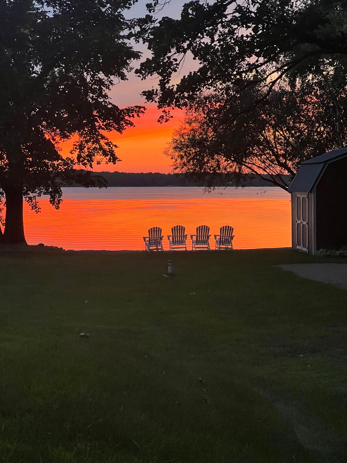 Four adirondack chairs are positioned in silhouette near the water's edge, facing a vibrant sunset that reflects off the lake. Tall trees frame the scene, enhancing the serene atmosphere of the lakeside setting.