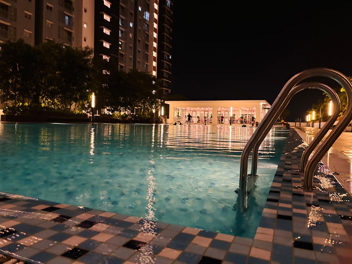 A swimming pool glimmers under the night sky, with gentle ripples reflecting light from surrounding fixtures. A ladder leads into the water, while a fitness center is visible in the background, creating a serene ambiance for relaxation.