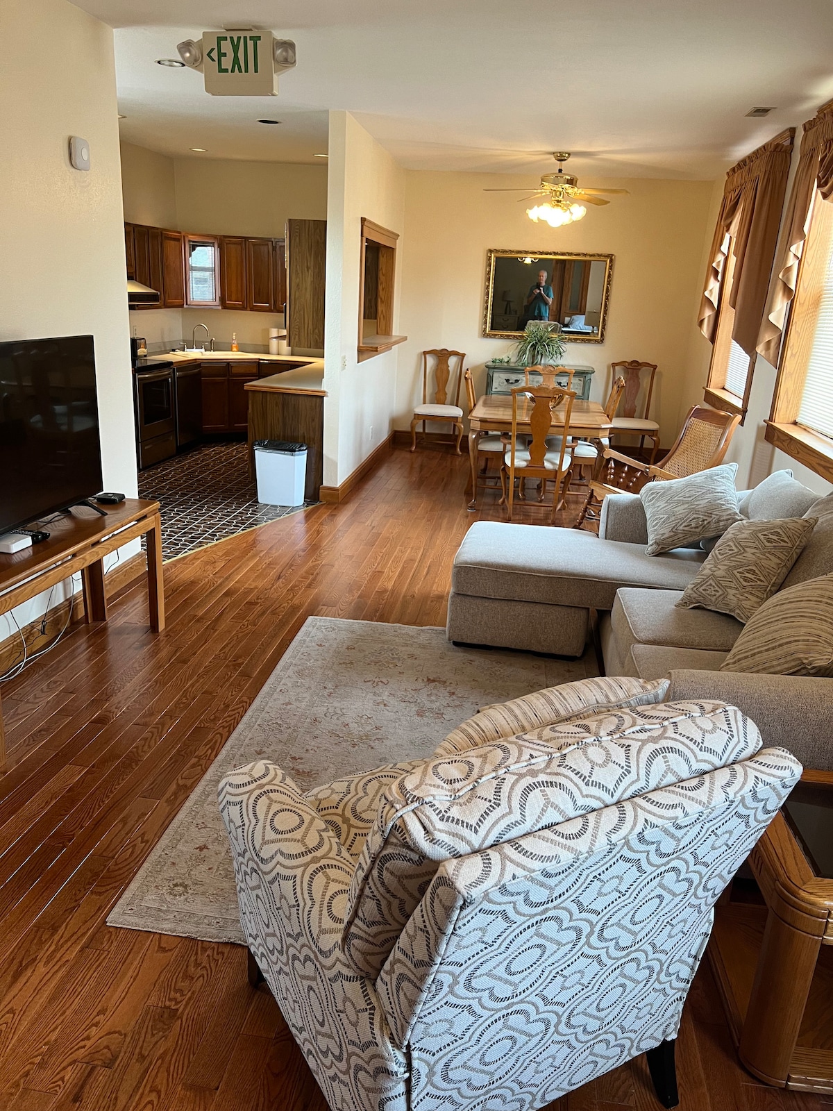 A living area showcases a comfortable sectional sofa and an accent chair, with a medium-sized television positioned against the wall. The dining space features a wooden table surrounded by four chairs, with a view into a well-equipped kitchen, illuminated by natural light from nearby windows.