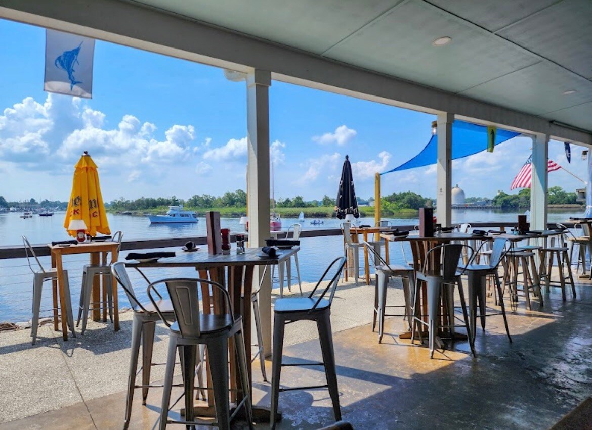 An outdoor dining area is showcased with metal tables and chairs, facing a scenic water view. Colorful umbrellas provide shade, and flags are seen gently waving in the breeze. The calm water reflects the blue sky, enhancing the serene atmosphere.