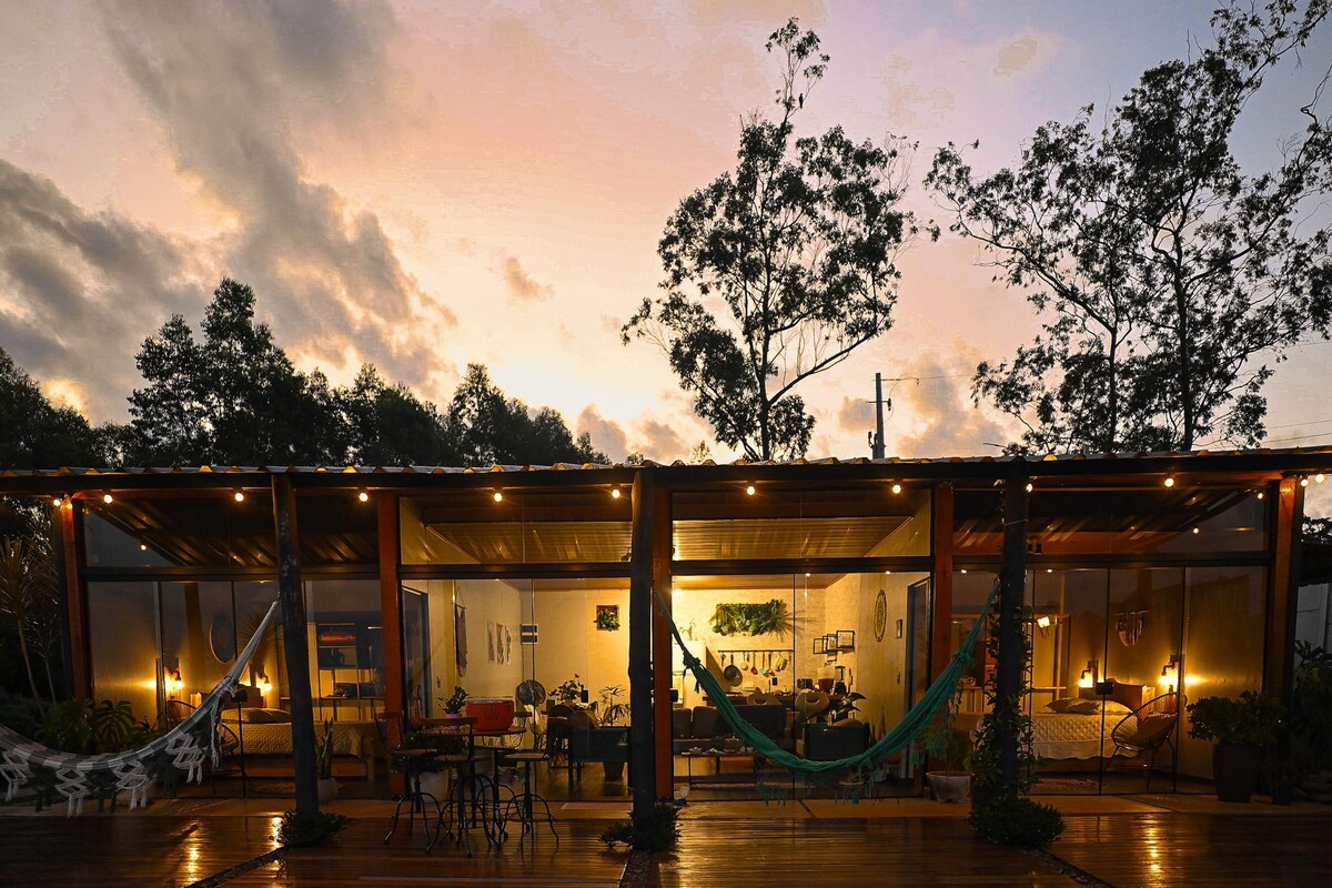 A charming house exterior is highlighted by warm lighting at dusk. The covered patio features hanging hammocks and a dining area, surrounded by lush greenery. Large windows provide a glimpse into the cozy interior while a serene sunset casts soft colors across the sky.