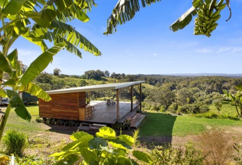 A spacious outdoor deck features wooden flooring and a covered area, overlooking lush greenery and rolling hills. Nearby, tropical plants provide a natural frame to the scene. The clear blue sky adds a serene backdrop to this tranquil retreat.
