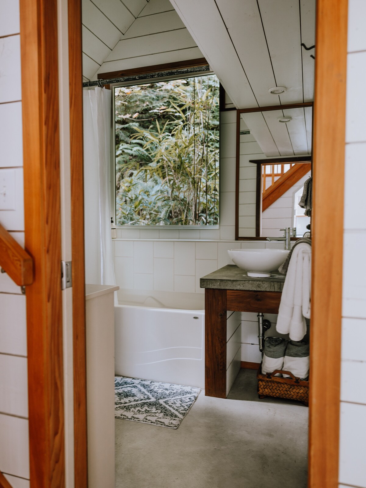 A modern bathroom is showcased, featuring a white bathtub and a wooden vanity with a round sink. Soft lighting enhances the bright, airy feel, while a window offers views of surrounding greenery. Towels are neatly arranged, adding to the organized appearance of the space.