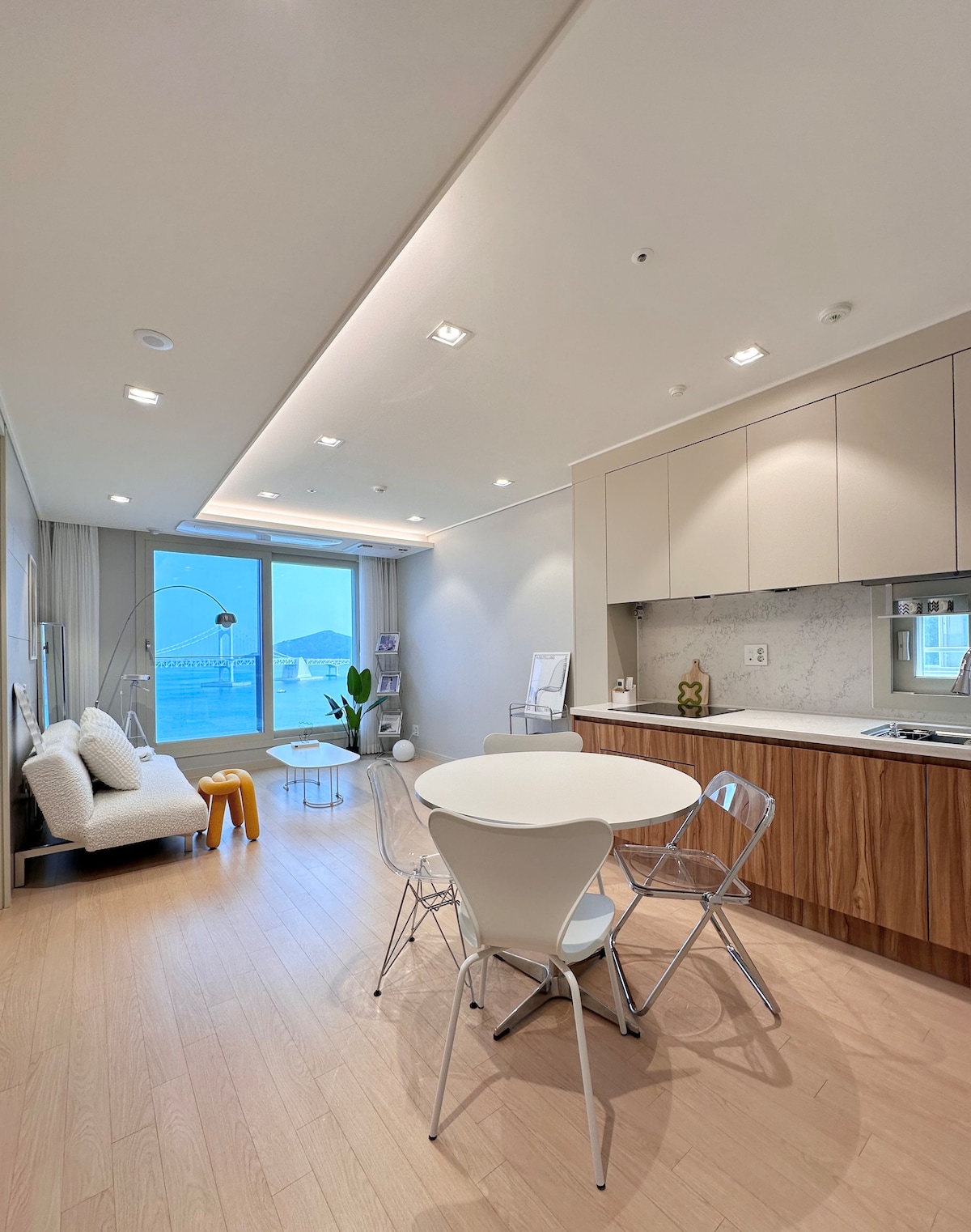 The modern living area features a dining table with four chairs, a plush sofa adorned with soft cushions, and a vibrant accent chair. Large windows allow natural light to fill the space, offering views of the surrounding area. Minimalistic cabinetry complements the light wooden flooring.