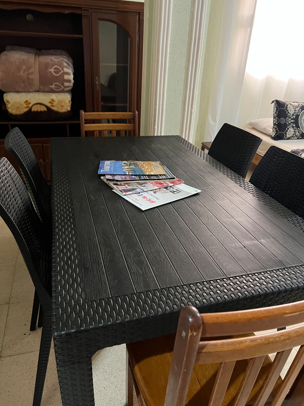 A dining table with a textured black surface is surrounded by six chairs. Magazines are neatly arranged on the table. A wooden cabinet in the background houses several items, while soft natural light filters through nearby window coverings.