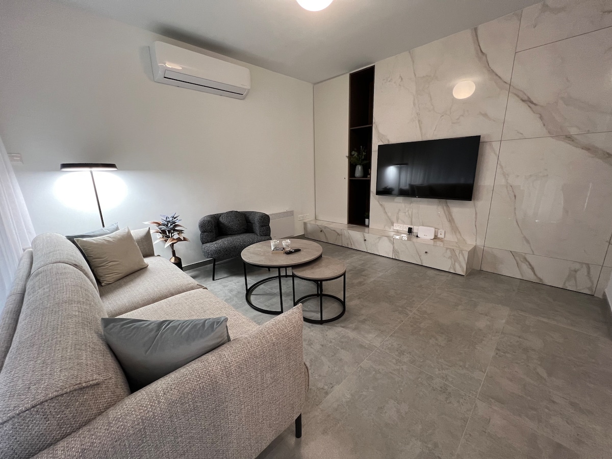 A modern living room showcases a comfortable sofa and two armchairs, arranged around a circular coffee table. A flat-screen TV is mounted on a marble-featured wall, while an air conditioning unit is visible above. Natural light filters through sheer curtains.