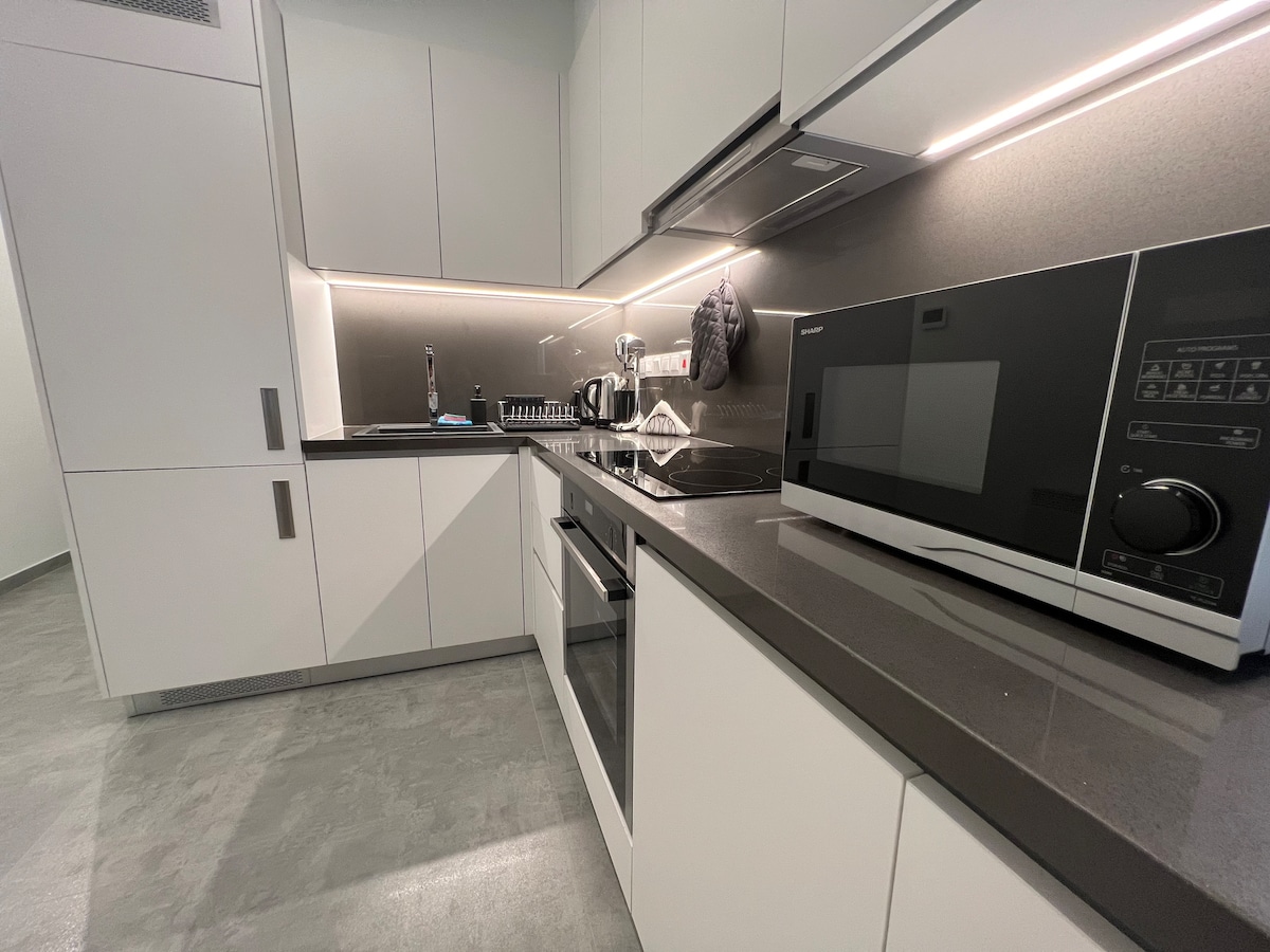 A modern kitchen is presented with sleek white cabinetry and dark countertops. Essential appliances include a microwave, toaster, coffee machine, and an oven. Ample countertop space is available for food preparation, with under-cabinet lighting enhancing visibility.
