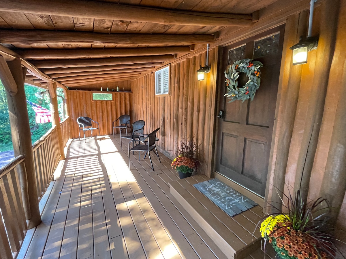 A rustic entryway features a wooden porch with a welcome mat and a wreath hanging on the door. Four metal chairs offer seating, with potted autumn flowers placed nearby. Natural light creates a warm ambiance along the wooden floor and walls.