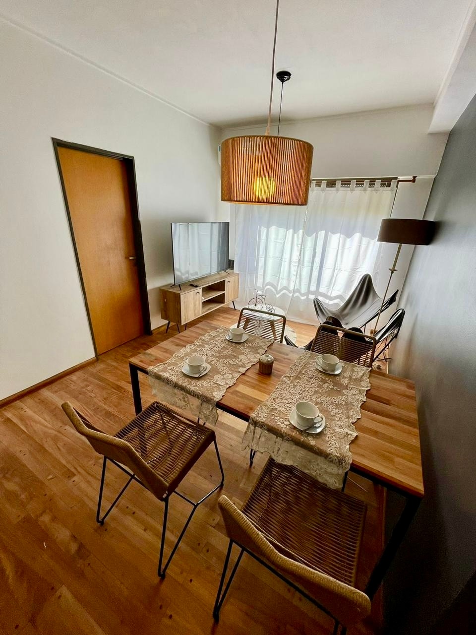 A dining area features a wooden table set for four, with woven chairs arranged around it. A pendant light illuminates the space, while a large window with sheer curtains allows natural light to fill the room. A television and a cozy armchair are also visible.