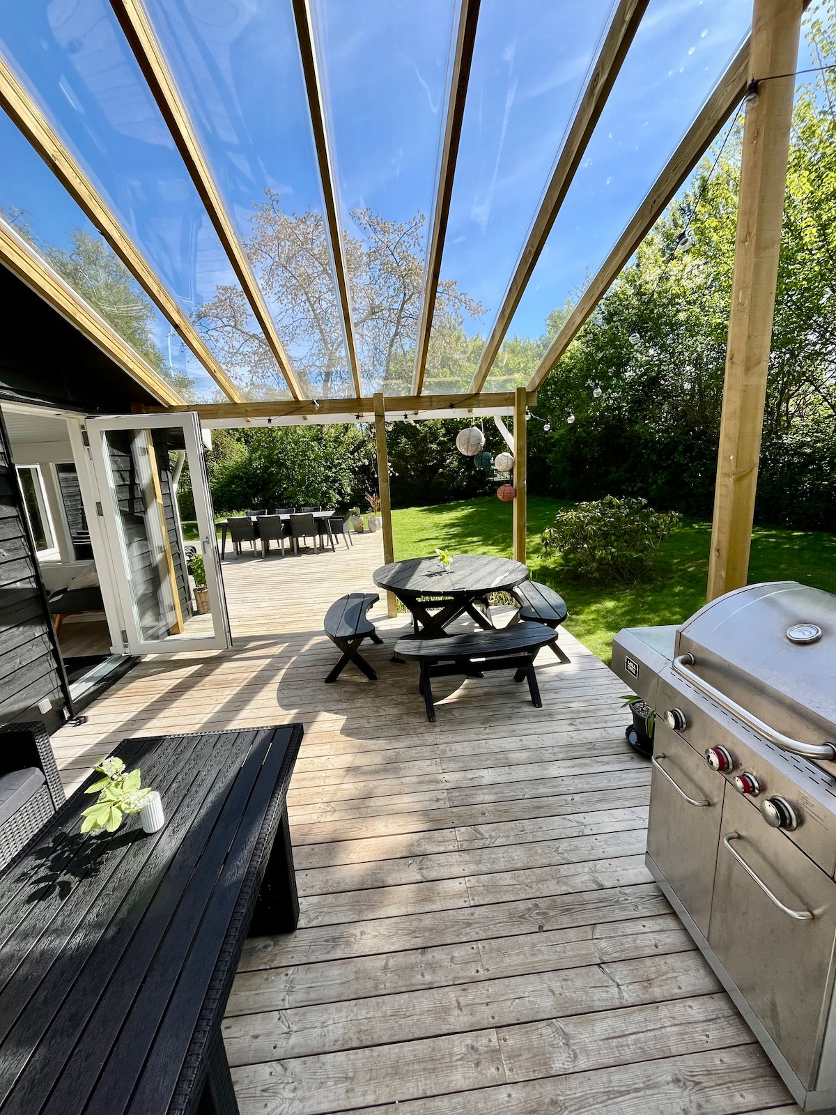 An outdoor terrace is featured, covered by a transparent roof, allowing natural light. A black picnic table and benches are positioned on wooden decking beside a gas grill. Lush greenery surrounds the area, creating a serene outdoor setting.
