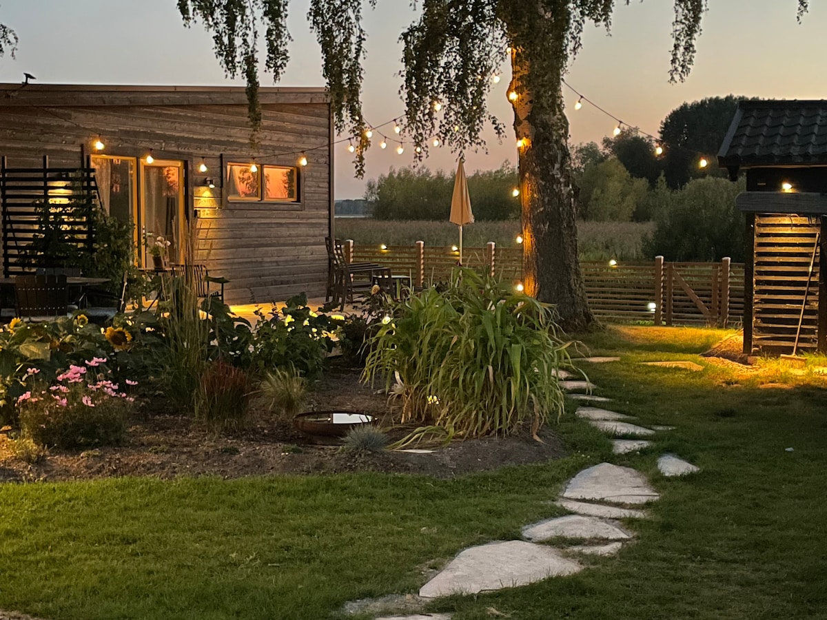 A charming outdoor space is illuminated by string lights as dusk settles. A stone pathway leads to a wooden guesthouse, surrounded by greenery and colorful flowers. Nearby, seating areas invite relaxation, while a patio umbrella offers shade within the intimate garden setting.