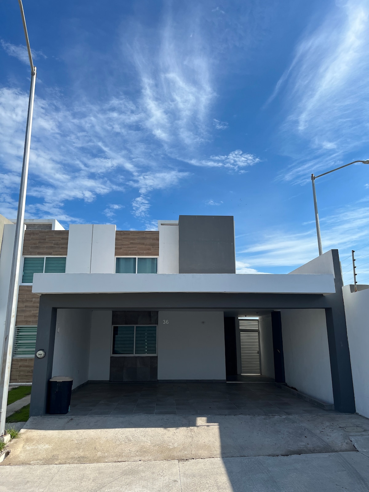 A modern two-story house is displayed under a clear blue sky, featuring a minimalistic facade with a mix of light and dark colors. The entrance is sheltered by a carport accommodating three vehicles, while a path leads to the door.