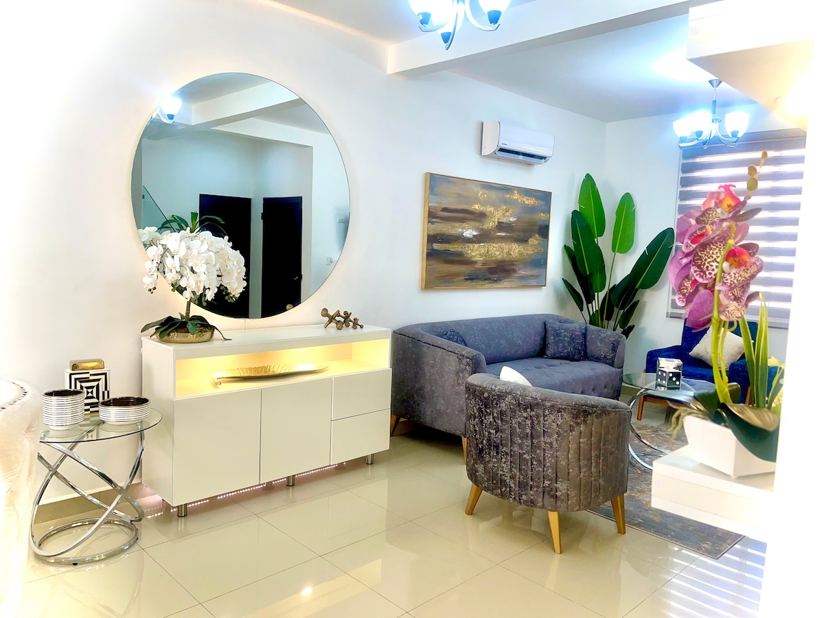 A stylish living area features a circular mirror above a modern sideboard with decorative accents. A cozy gray couch is positioned beside an indoor plant, with unique art displayed on the wall. Bright lighting from the ceiling complements the inviting atmosphere.