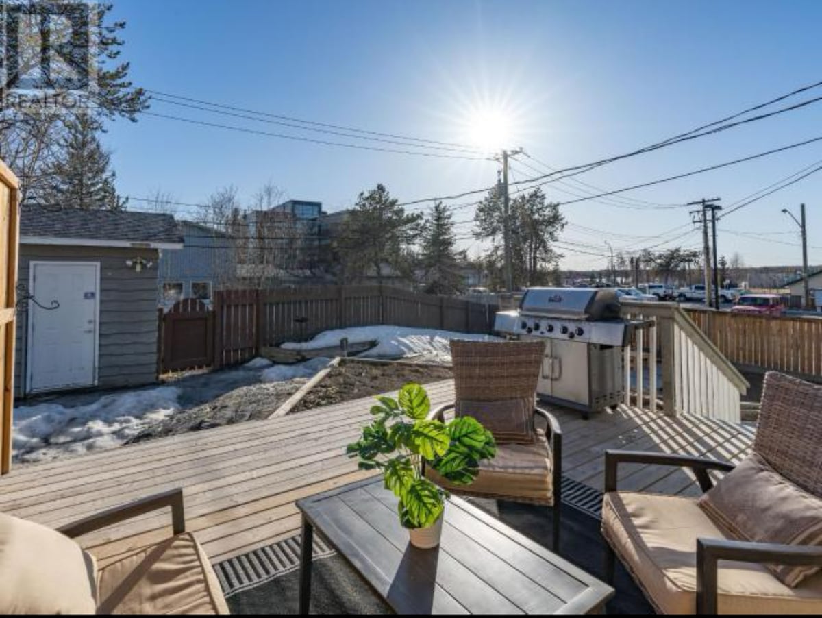 An outdoor deck area is presented, featuring two comfortable chairs alongside a table. A barbecue grill is visible, with greenery around. Sunlight casts a warm glow over the space, highlighting the clear blue sky and the nearby fence, while remnants of snow remain in the background.