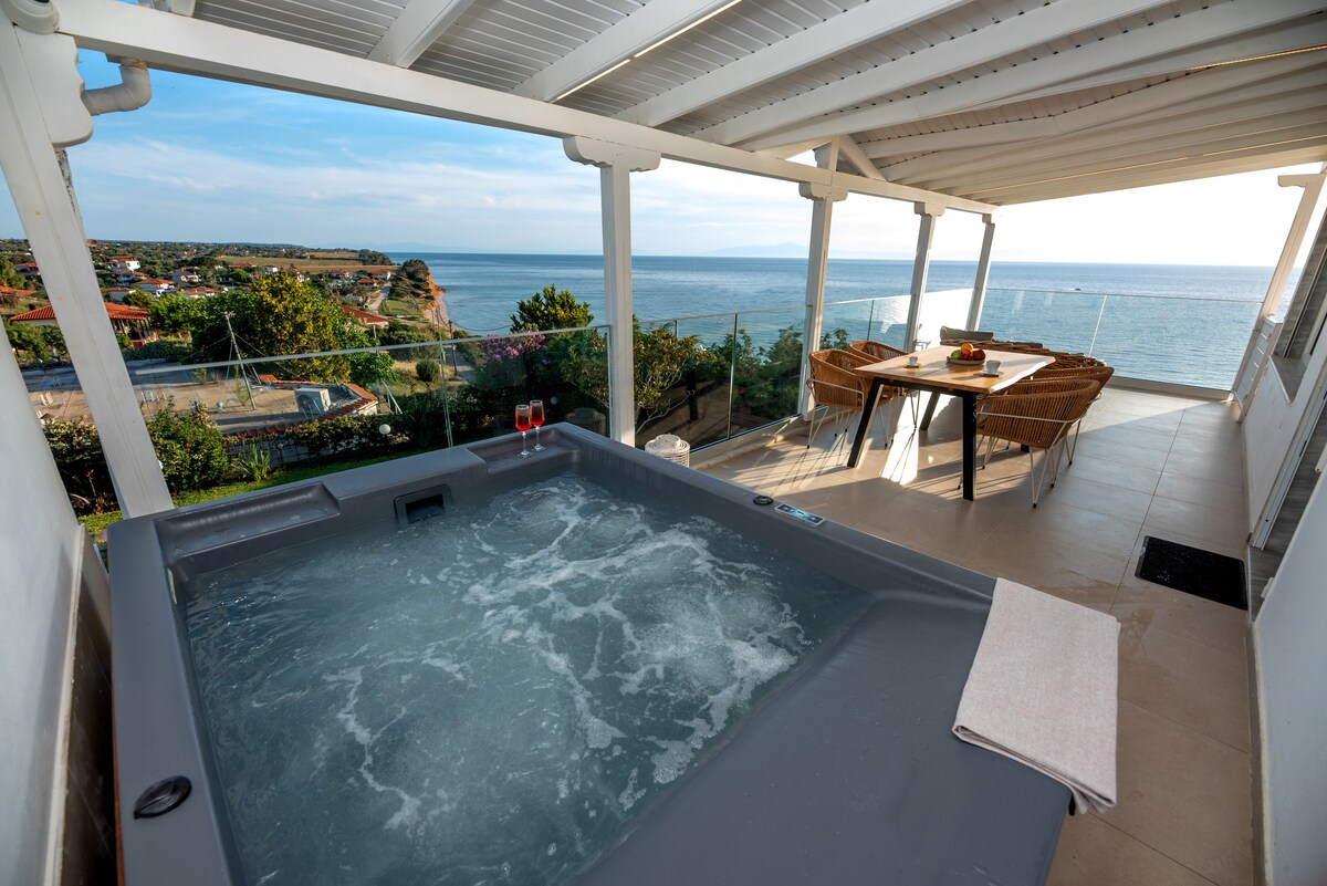 A private jacuzzi is situated on a terrace overlooking the Aegean Sea. The scene includes a dining area with woven chairs and a table, providing a scenic view of the horizon through glass railings. Lush greenery frames the space, enhancing the tranquil ambiance.