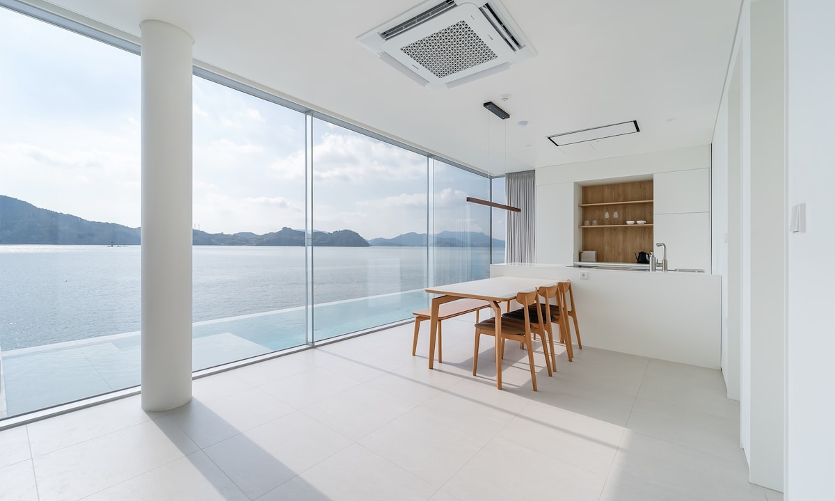 A bright dining area is illuminated by natural light, featuring a minimalist table with wooden chairs positioned by expansive glass windows. The serene ocean view is visible in the background, enhancing the spacious feel of the room.