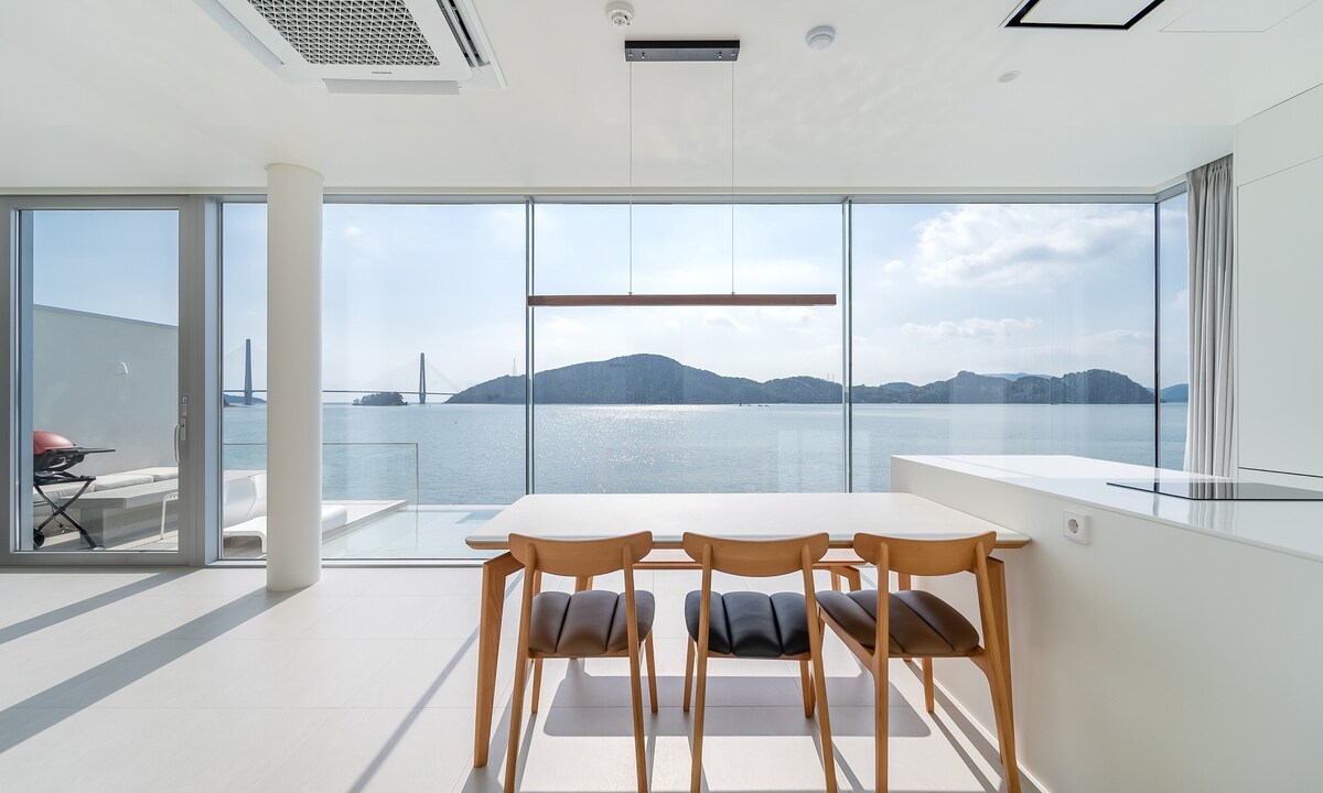 An open dining area features a wooden table with seating for three, positioned near large glass windows that provide views of the ocean and surrounding mountains. Natural light floods the space, enhancing the minimalist design and modern furnishings.