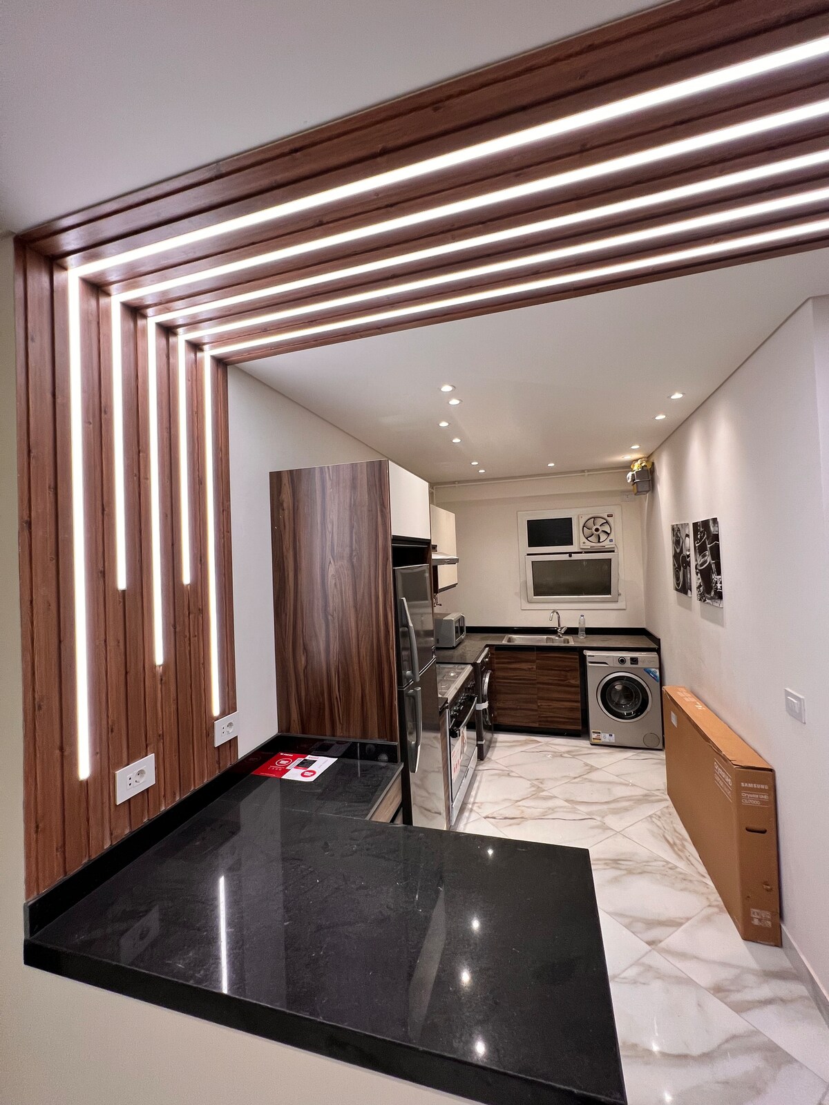 The compact kitchen features a dark granite countertop and modern cabinetry with wood accents. Stainless steel appliances include a fridge, oven, and washer. Soft lighting is provided by linear fixtures along the wall, creating a bright and functional cooking space.