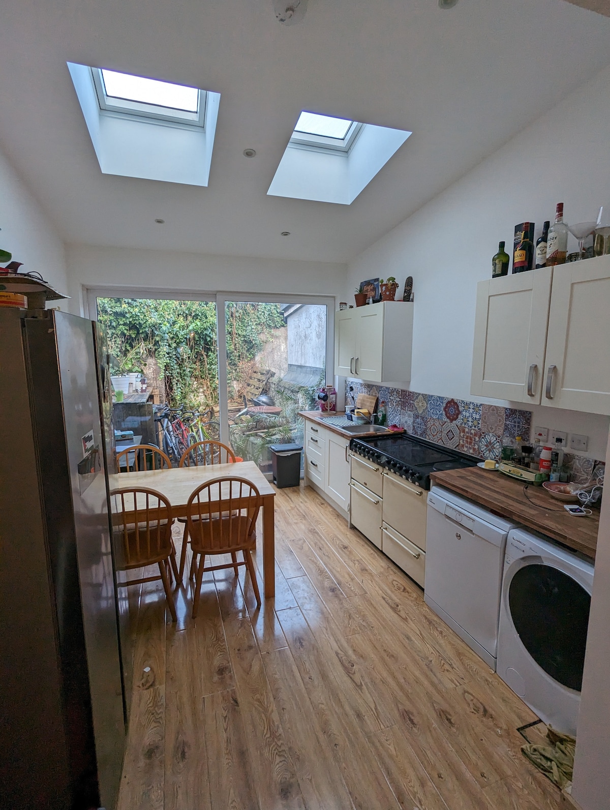 A bright kitchen features skylights allowing natural light to fill the space. A dining table with four wooden chairs is situated next to a range of cabinets and appliances. Large glass doors open to a garden area, enhancing the indoor-outdoor connection.