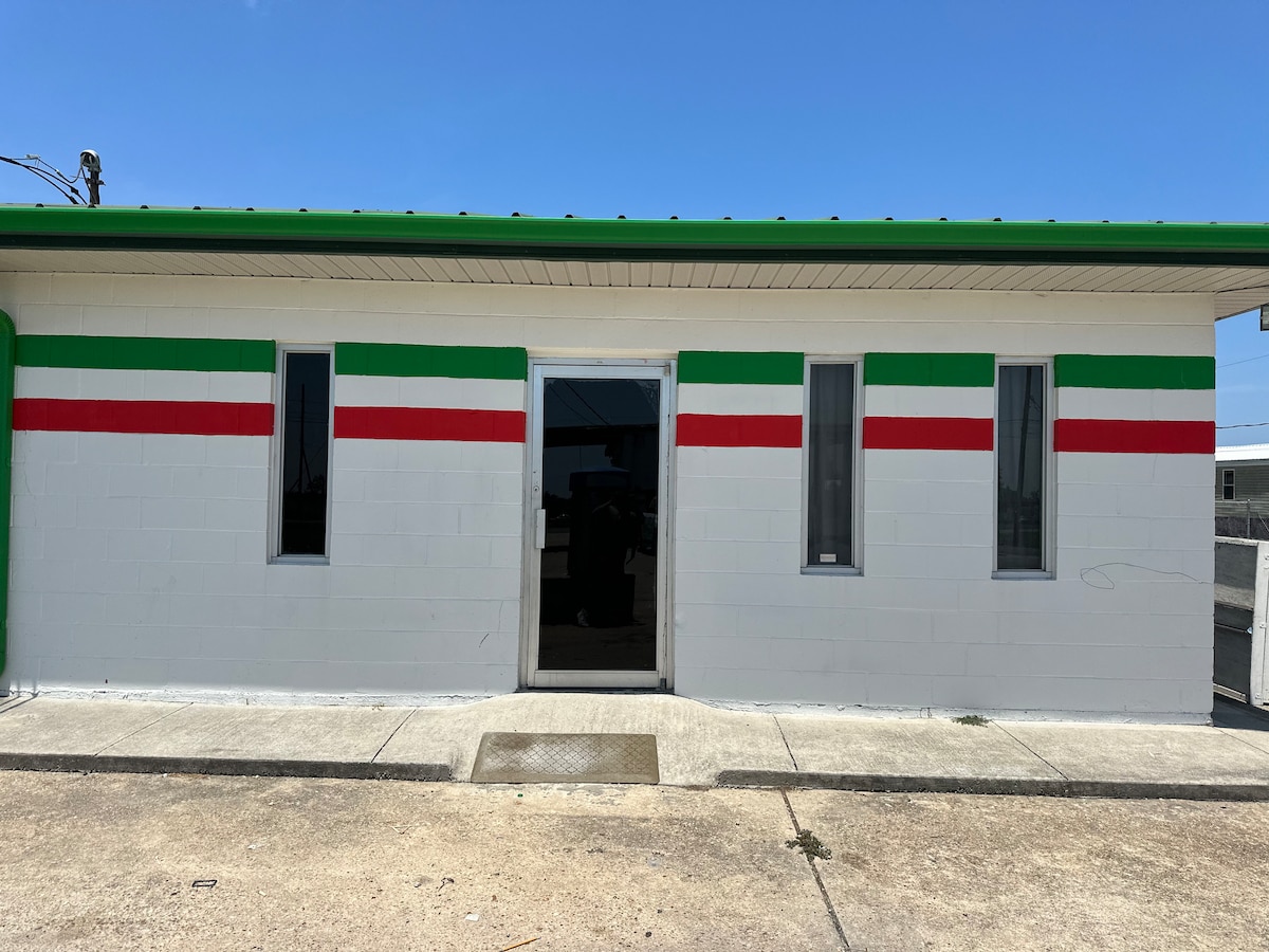 The exterior of a freshly remodeled apartment is showcased, featuring a modern design with distinct horizontal stripes of green and red on a white wall. Two large windows on either side of the central entrance reflect natural light, adding to the inviting appearance.