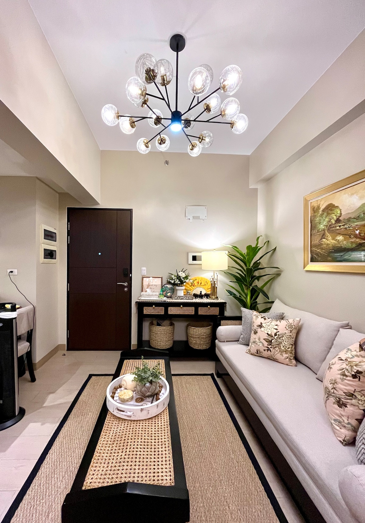 A stylish living area is presented with a soft gray sofa adorned with decorative pillows. A modern chandelier illuminates the space, while a decorative coffee table features a tray with plants and decorative items. A slender indoor plant adds a touch of nature.