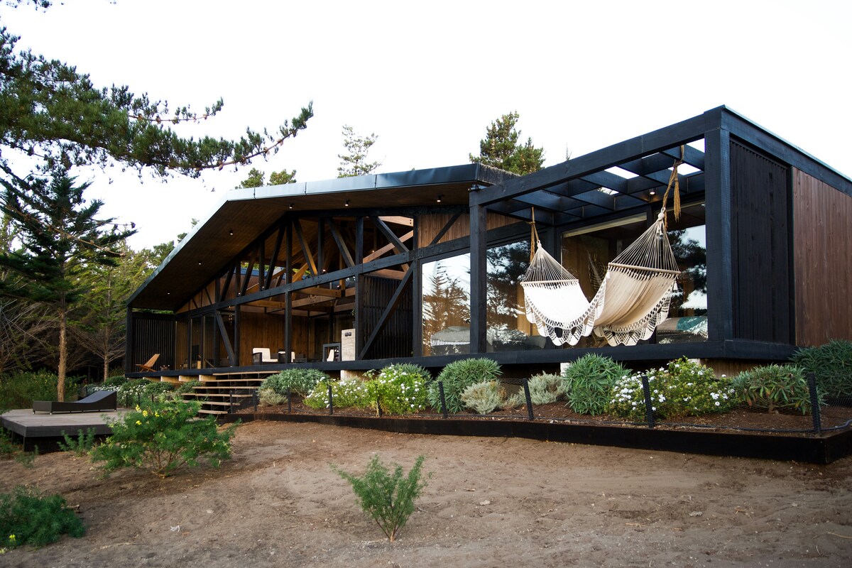 A modern house is seen nestled among trees, featuring a large deck with two hanging hammocks. The structure has large windows that integrate with the surrounding nature and a stylish wooden design that complements the outdoor landscape.