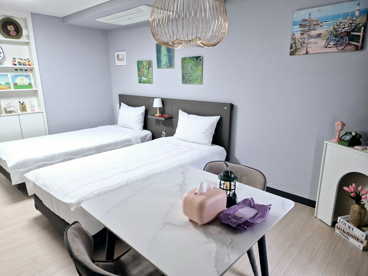 A comfortable room features two spacious beds with white linens, complemented by a modern table and chairs. Soft lighting is provided by a decorative lamp. Wall art and shelves with decorative items add character to the space.