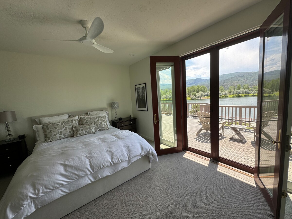 A serene bedroom features a spacious bed with white bedding, complemented by two nightstands with lamps. Large glass doors open to a deck, offering views of a lake and mountains, while natural light fills the room. A ceiling fan provides additional comfort.