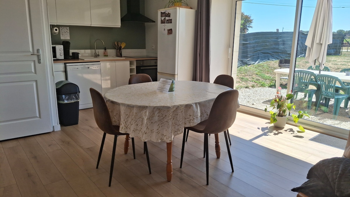 A dining area is presented with a round table surrounded by four chairs. Natural light fills the space from a large sliding glass door, which offers a view of the outdoor seating area. A kitchen area is visible in the background.