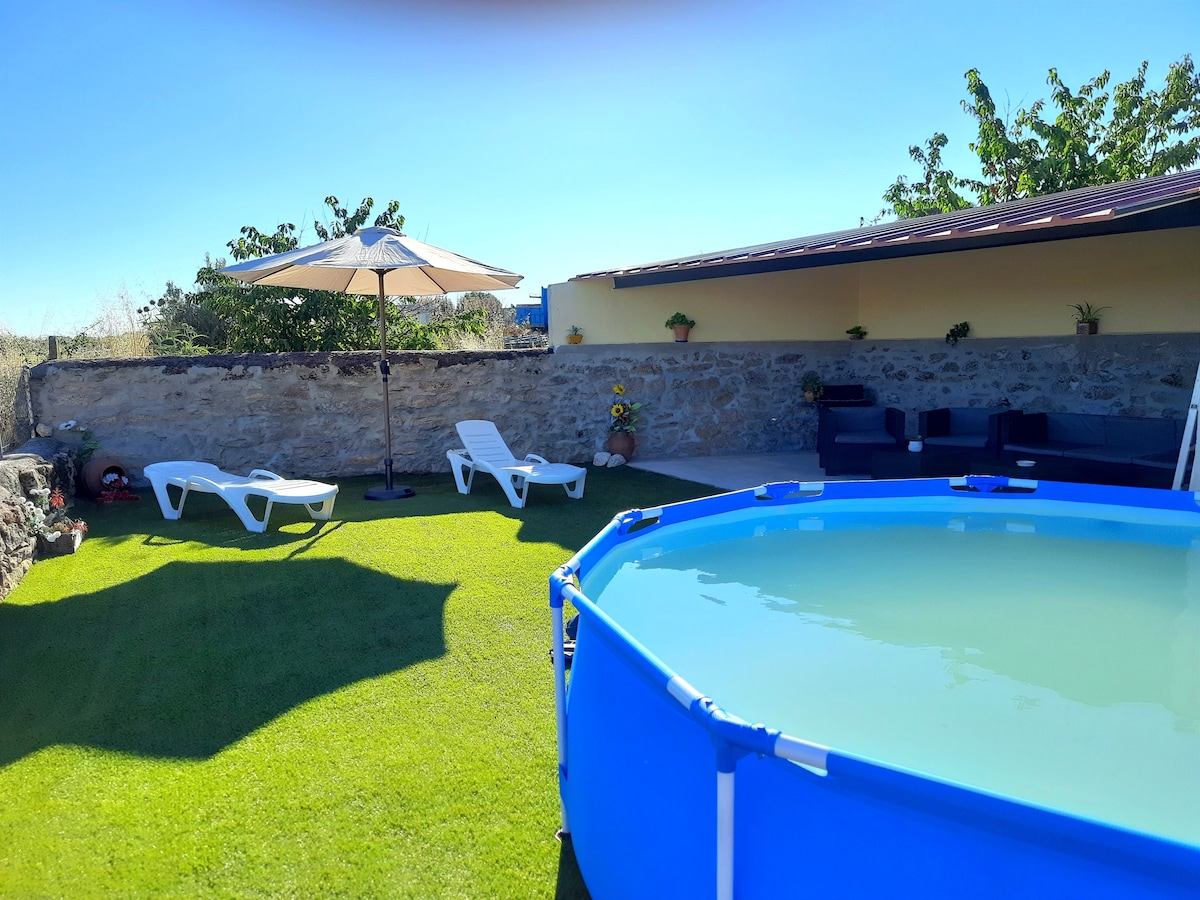 An outdoor space is presented featuring a circular pool surrounded by manicured grass. Two white lounge chairs and a shaded seating area with sofas provide relaxation options. Sunlight illuminates the scene, enhancing the greenery and the tranquility of the environment.