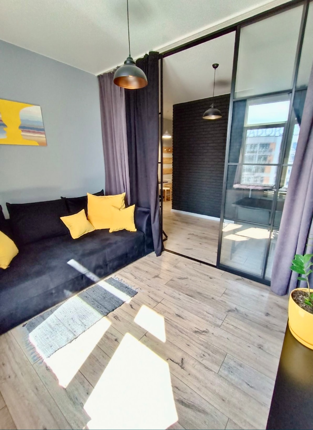 A bright living area features a dark sofa adorned with yellow pillows, inviting relaxation. Natural light streams through the large glass doors, illuminating the light-toned wooden floor. A potted plant and modern light fixtures add subtle style to the room, separated by elegant curtains.