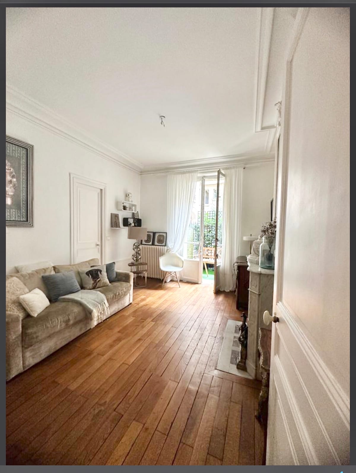 A bright living space is highlighted by natural light filtering through tall windows. A cozy sofa is positioned against the wall, complemented by decorative pillows. The warm, wooden flooring adds a classic touch. Views of an inner garden are visible through the glass doors.