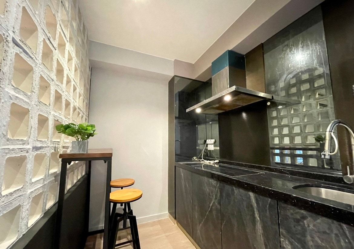 An open kitchen is presented with sleek black cabinetry and marble countertops. Two wooden bar stools are positioned at a small bar table. A wall of glass blocks allows natural light to enter while providing a modern aesthetic.