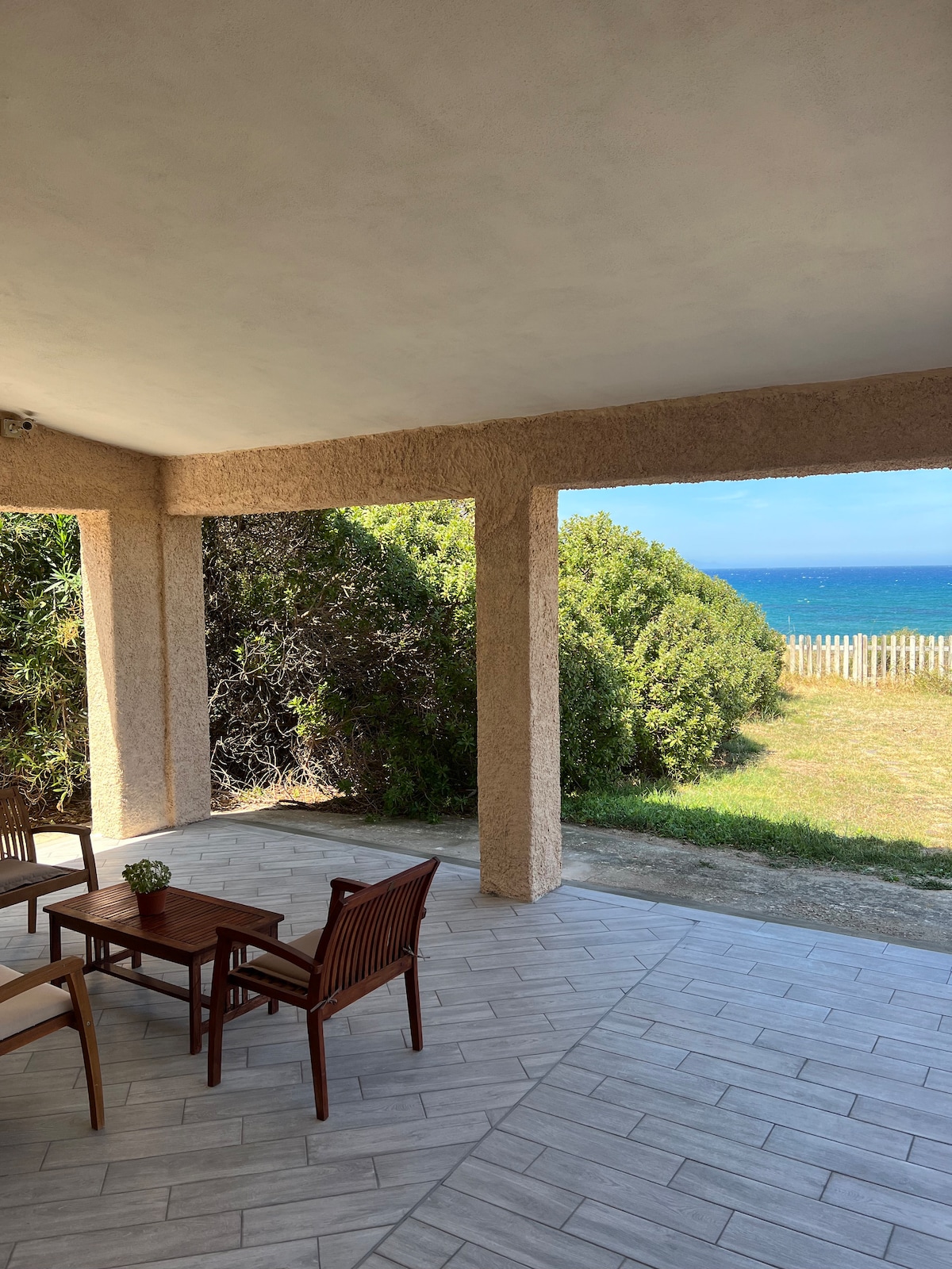 A covered outdoor area features a wooden table accompanied by four chairs, providing a suitable space for relaxation. Lush greenery borders the view, leading to the expansive blue sea, visible beyond a grassy area and wooden fence.