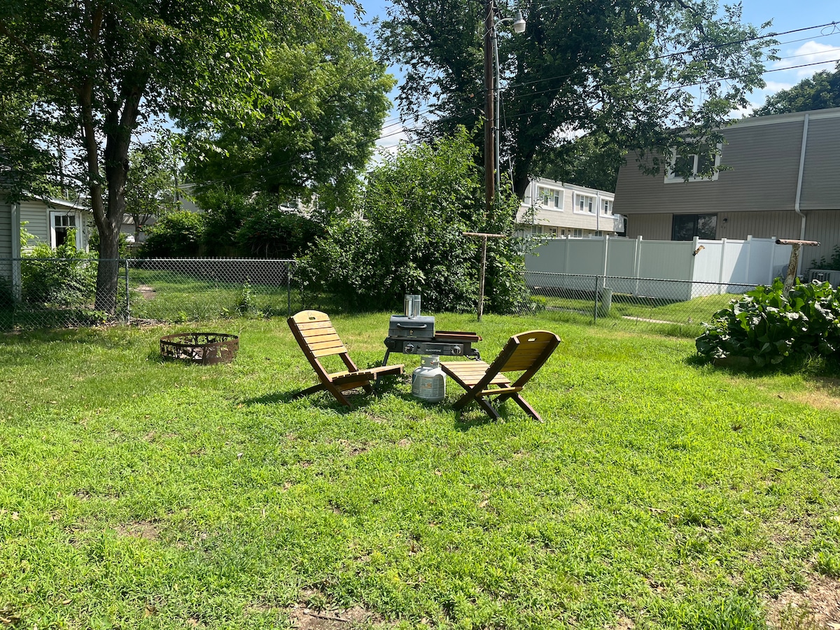 A grassy yard features two wooden chairs positioned around a small circular table, providing a cozy seating area. A fire pit is located nearby, surrounded by pebbles. Lush greenery and trees create a natural backdrop, enhancing the outdoor space's relaxed atmosphere.