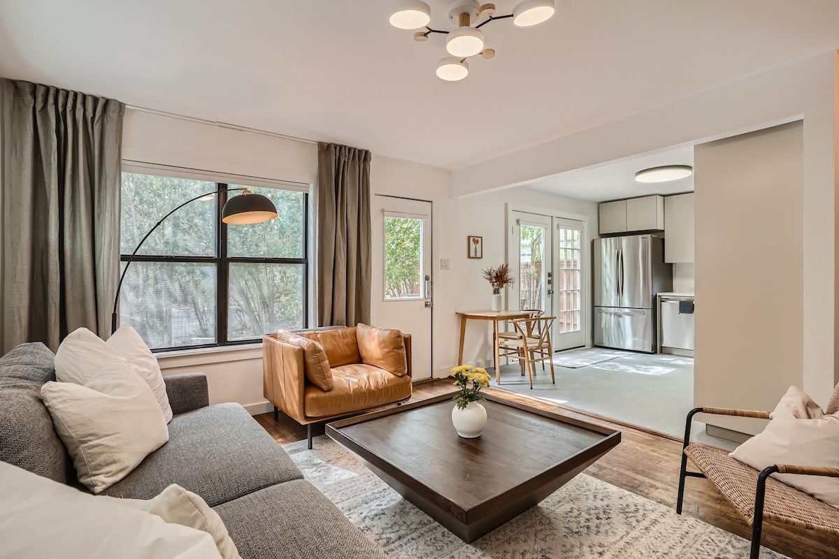 A stylish living area features a gray sofa and a leather chair, complemented by a large wooden coffee table. Natural light filters through a wide window adorned with light curtains. The open layout leads to a dining space visible in the background.