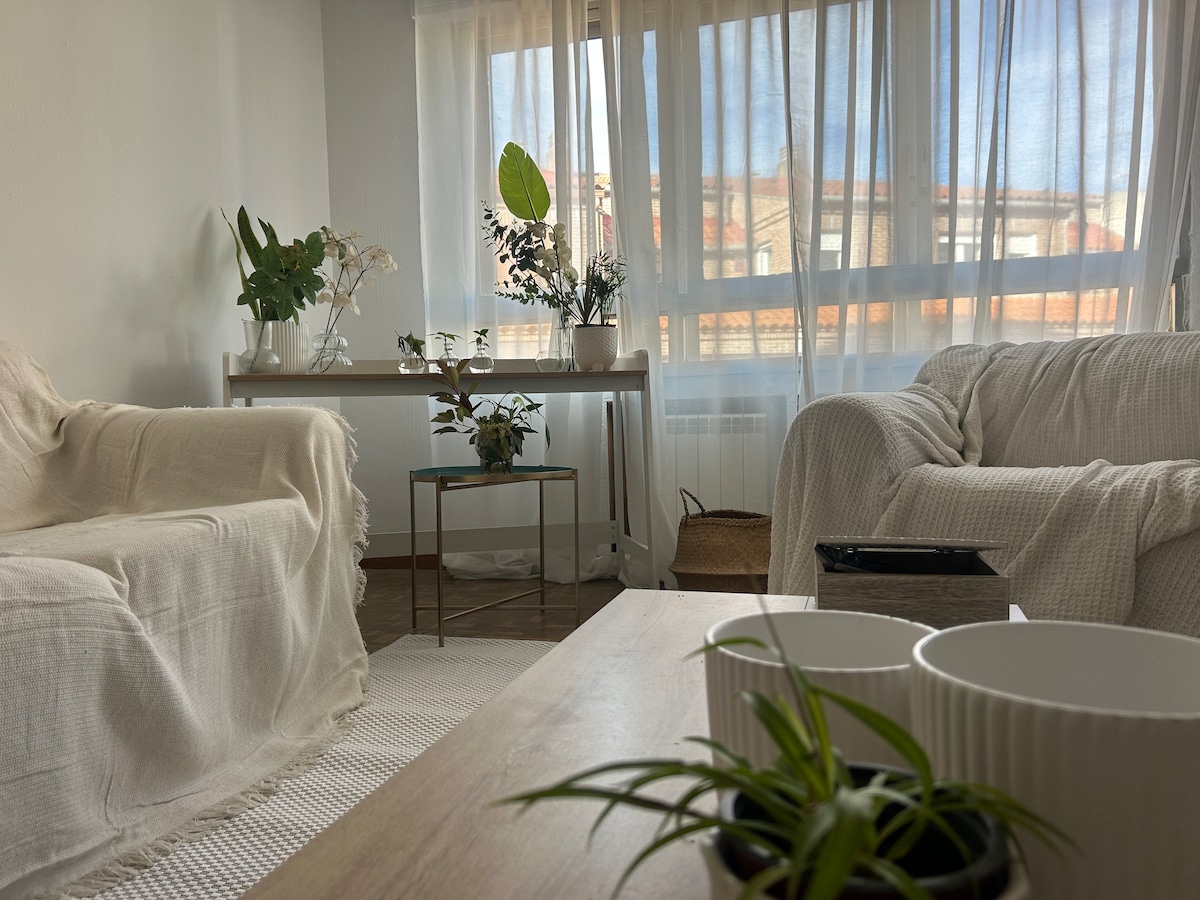 A bright living area is filled with natural light, highlighting neutral-colored sofas draped in light fabric. A wooden coffee table sits in the foreground, adorned with potted plants and decorative items. Large sheer curtains frame the window, creating a soft, airy ambiance.