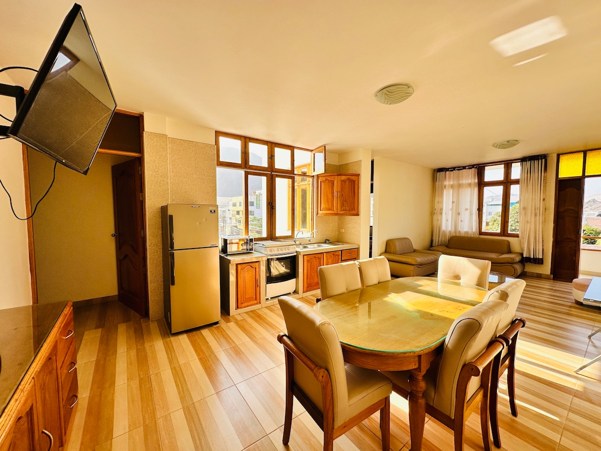 A dining area is highlighted by a large circular table surrounded by six chairs. An adjacent kitchen features wooden cabinetry and a stainless steel refrigerator. Natural light enters through large windows, illuminating the spacious room with light-colored wood flooring.